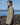 Side view of a person wearing a beige waterproof jacket, standing on a rocky shoreline with the sea and hills in the background.