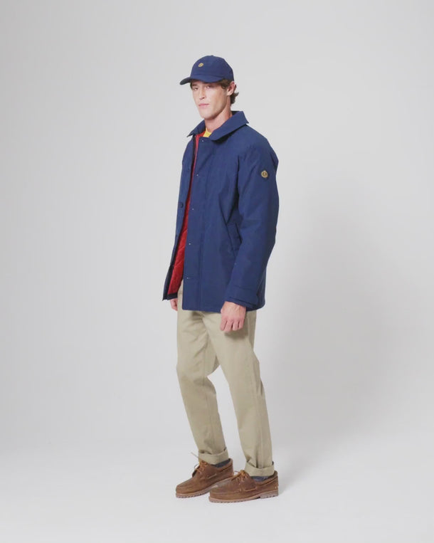 Person wearing a navy waterproof jacket, beige trousers, brown shoes, and a navy cap, standing against a white background.