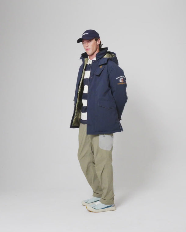 Waterproof jacket styled over a striped rugby shirt and trousers, paired with a cap, photographed in studio.